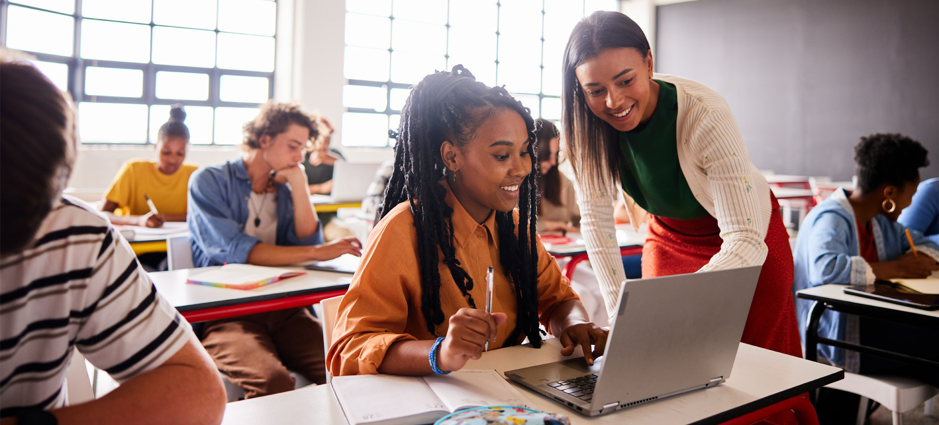 Smiling teacher talking with a student using a laptop during a classroom lesson